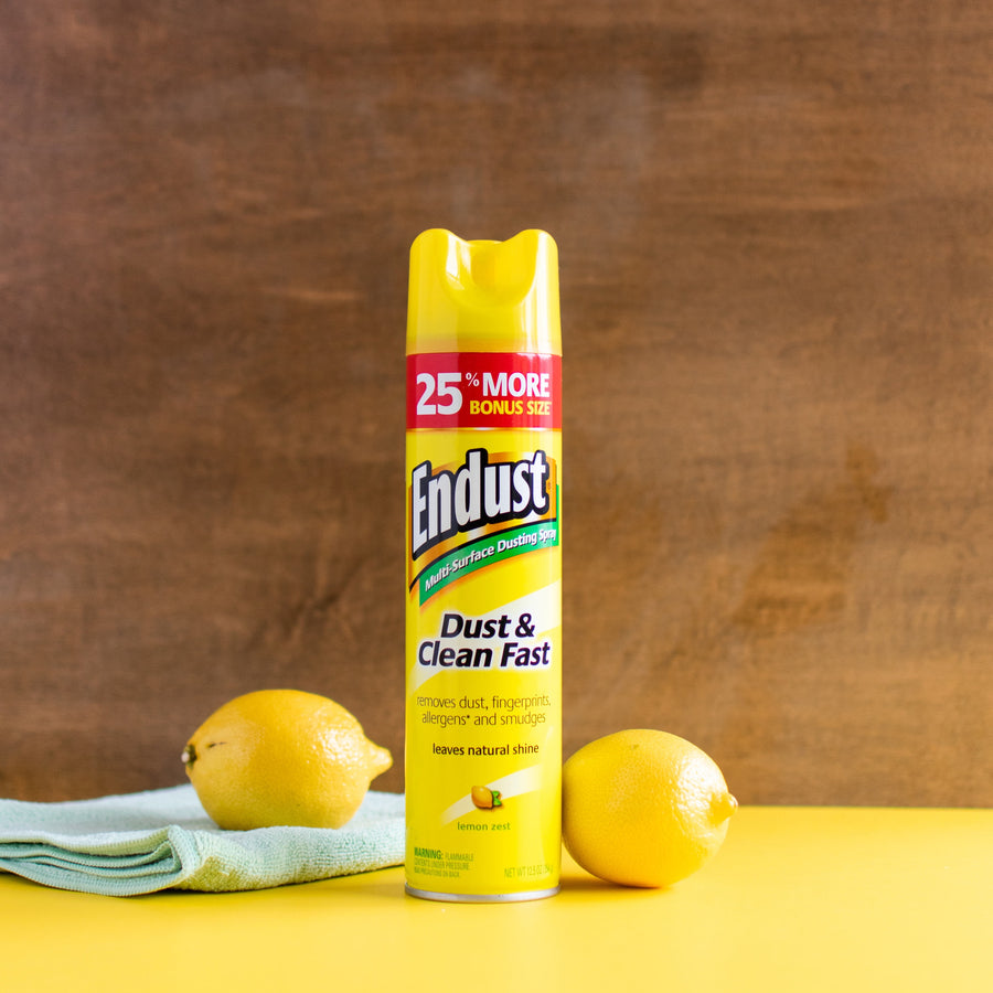 Centered Endust lemon zest aerosol can standing on a yellow surface with lemons and a cloth against a wood backdrop