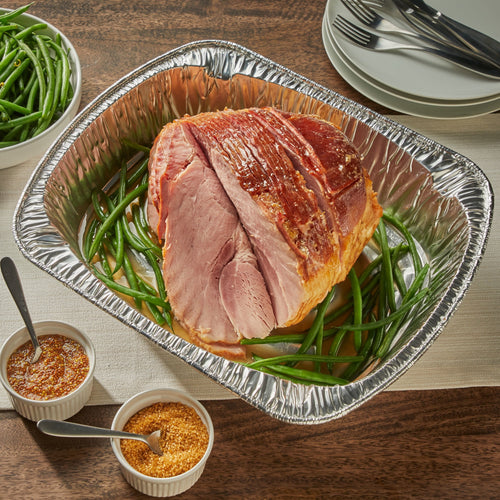 Disposable aluminum roaster pan holding a glazed ham on bed of green beans with serving dishes nearby.