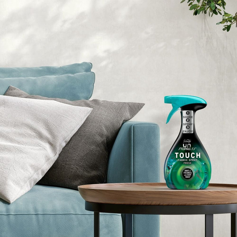 Febreze Unstopables Touch fabric spray bottle shown on a wooden table beside a sofa in a living room setting.