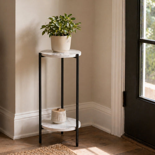 Two-tier marble-top plant stand with black metal legs styled in a corner with a potted plant on top and a small ceramic piece below