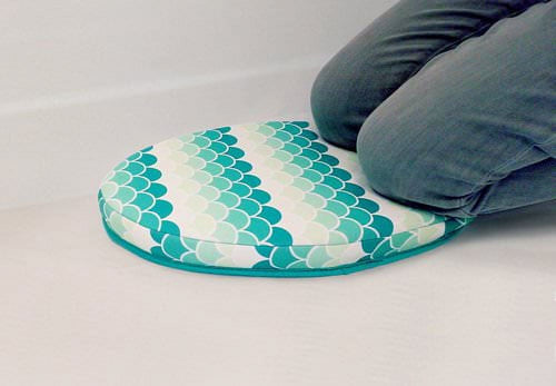 Teal and white scallop-pattern oval bath kneeler pad with someone kneeling on it, low studio background.