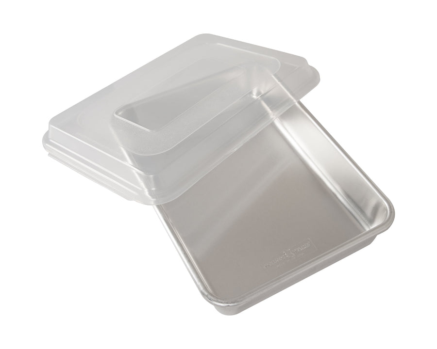 Aluminum rectangular baking pan with a clear plastic storage lid partially removed, on white background