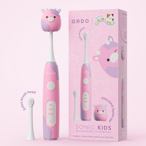 Pink Ordo kids sonic toothbrush with cow-themed Squishmallows cap shown next to its matching pink product box and extra brush head.