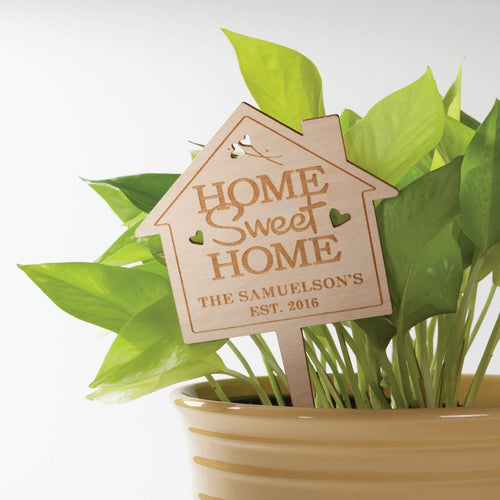 Laser-engraved wooden house-shaped plant stake reading “HOME Sweet HOME” with a personalized name, placed in a potted green plant.