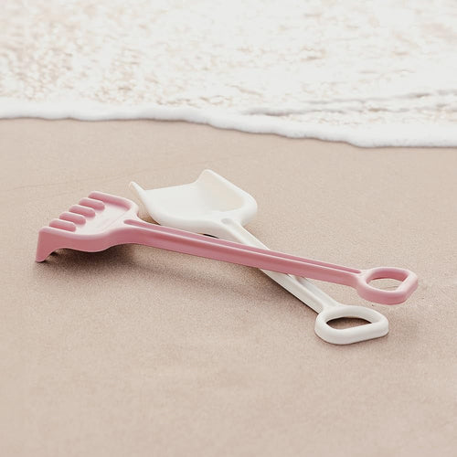 Pink toy rake and cream spade placed on wet sand at the waterline on a beach