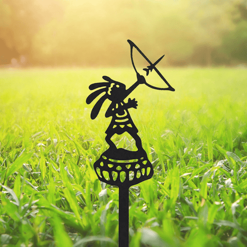 Black metal garden stake shaped like a tribal figure holding a curved umbrella, set in bright green grass.