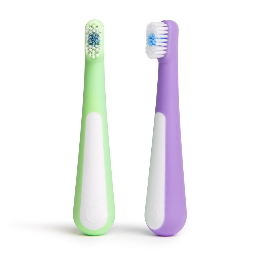 Two Ubbi toddler toothbrushes standing upright on white background, one purple and one green with white grip panels.