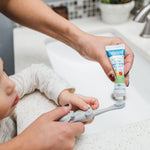 Adult squeezing Dr. Brown's baby toothpaste onto a gray otter toothbrush for child