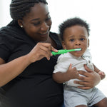 Adult brushing a baby’s teeth using a green crocodile toddler toothbrush in a warm, natural lifestyle setting.