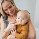 Smiling adult holding a baby while using a yellow silicone finger toothbrush in the baby's mouth