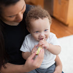 Adult holding a toddler on their lap while brushing the child's teeth with a small toothbrush in a home setting.