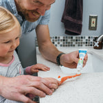 Adult and child at a bathroom sink applying toothpaste to an orange Dr. Brown's toddler toothbrush in lifestyle scene.