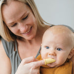 Adult holding a baby while using a yellow silicone finger toothbrush in the infant's mouth in a close lifestyle shot