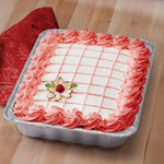 Baked frosted square cake in an aluminum foil pan on a wooden surface with a red patterned napkin