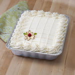 Iced square cake presented in an aluminum foil pan on a wooden table with a green patterned napkin