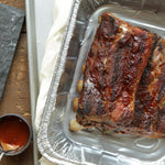 Close overhead view of barbecued ribs placed in a disposable aluminum roasting pan on a baking sheet
