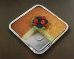Baked square cake in a foil aluminum pan with a slice removed and topped with berries and herbs on a dark surface.