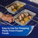 Two aluminum foil trays with cooked meat and potatoes on oven racks, hand reaching to one tray