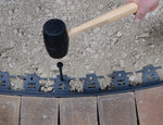 Lifestyle image showing a mallet driving a black nylon anchor spike into garden edging installed in soil.
