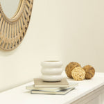 Angled lifestyle shot of a small white bubble ceramic planter on books with decorative woven balls and a mirror at left.