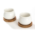 Two white jar-shaped ceramic pots shown from an angled top view each seated on a round bamboo tray on white background