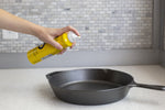 Hand holding a can of Lodge seasoning spray being applied toward an empty cast iron skillet on a kitchen countertop.