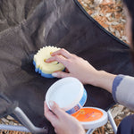 Person applying a sponge to a dark fabric chair seat beside an open tub of PowErase Gel on the armrest