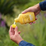 Two hands pouring liquid from a yellow Arber bottle into its measuring cap against a blurred garden backdrop