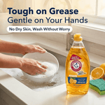 Hands washing a dish at a sink with an Arm & Hammer + OxiClean Fresh Citrus bottle and sponge visible at the edge.