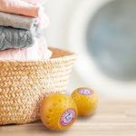 Lifestyle scene with two orange Arm & Hammer Odor Busterz balls beside a woven laundry basket and folded clothes.
