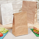 Three self-standing paper lunch bags in brown and white displayed on a wooden board with sandwich ingredients nearby.