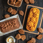 Styling shot of two patterned loaves and assorted leaf-shaped cookies on dark table, one loaf with pumpkin motif