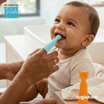 Adult finger using a blue silicone finger brush in a smiling baby's mouth, with an orange brush standing nearby and Shark Tank badge visible.