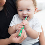 Baby biting a green crocodile-shaped toddler toothbrush while an adult supports the child, bright soft-lit lifestyle image.