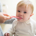 Baby being gently brushed with a pink flamingo-shaped toddler toothbrush held by an adult in a bright bathroom scene