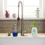 Kitchen sink styled lifestyle shot showing a Babyganics foaming dish soap bottle with green pump placed beside a faucet on a countertop.