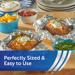Baked potatoes topped with sour cream, cheese and chives resting on crumpled aluminum foil with bowls of toppings nearby