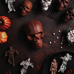Baked skull-shaped cakes and smaller spooky treats arranged on a dark surface with pumpkins and skeleton-shaped confections.