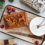 Baked holiday loaf with Santa sleigh and reindeer design on a wooden tray beside the Nordic Ware loaf pan