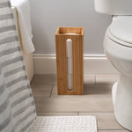 Lifestyle bathroom scene with bamboo toilet paper reserve on the floor between a toilet and a towel-covered hamper