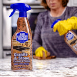 Bar Keepers Friend Granite & Stone bottle in foreground with person wearing yellow gloves wiping a countertop in the blurred background.
