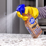 Hand wearing yellow glove spraying Bar Keepers Friend Granite & Stone cleaner onto a granite countertop, spray mist visible.