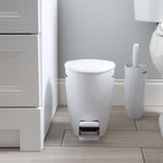 White pedal trash bin and matching toilet brush holder placed on a bathroom floor beside a toilet and vanity