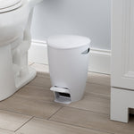 White pedal trash bin positioned on a bathroom floor next to a toilet and vanity baseboard in a lifestyle shot