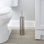 Chrome toilet brush holder placed on a bathroom floor beside a toilet and vanity