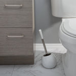 White dome toilet brush holder with brush standing between a cabinet and a toilet on tiled floor