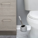 White rounded toilet brush holder with brush placed beside a toilet and gray cabinet on tiled floor
