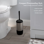 Matte black-handled toilet brush standing in its stainless-steel holder on a bathroom floor beside a toilet and cabinet, lifestyle shot.