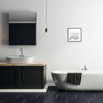 Minimal modern bathroom with a framed black-and-white toilet-paper quote print hanging on the wall above a freestanding tub.