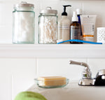 Bathroom shelf scene with glass jars, toiletries and a blue toothbrush lying across the ledge near a sink faucet